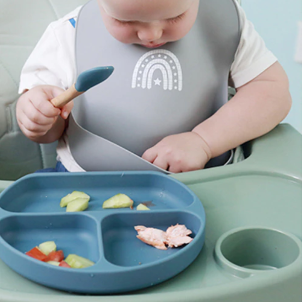 Baby With Silicone Bib Eating From Silicone Plate With Spoon On Highchair