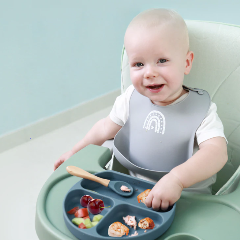Smiling Baby With Silicone Bib Eating From Silicone Plate With Spoon On Highchair